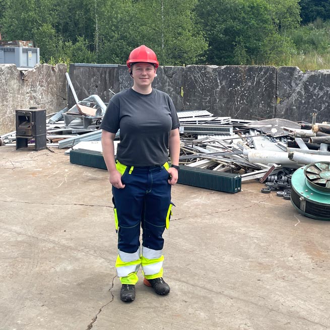 Sisse Bæk, skrotspecialist, kvinde stående i gult arbejdstøj med rød hjelm foran skrotdynge Sisse Bæk, skrotspecialist, kvinde stående i gult arbejdstøj med rød hjelm foran skrotdynge