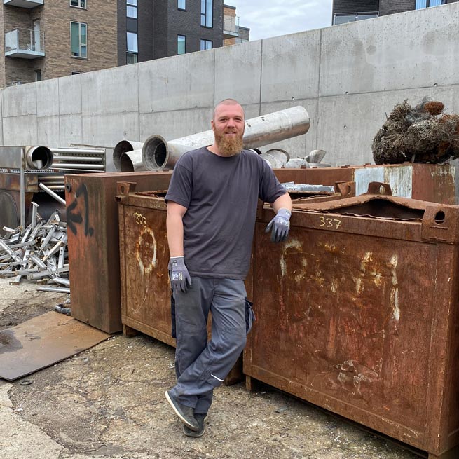 Marc fra Skrotspecialisten Odense står i gråt tøj foran brun container fyldt med gammelt jernskrot. Marc fra Skrotspecialisten Odense står i gråt tøj foran brun container fyldt med gammelt jernskrot.