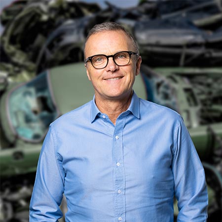 Portrætbillede af Kurt Søndergaard der er segmentchef for autoophug hos HJHansen. Han er iført blå skjorte og briller. I baggrunden er der biler i en bunke sendt til autoophug.