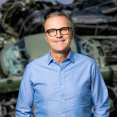 Kurt-Soendergaard-HJHansen-Recycling Portrætbillede af Kurt Søndergaard der er segmentchef for autoophug hos HJHansen. Han er iført blå skjorte og briller. I baggrunden er der biler i en bunke sendt til autoophug.